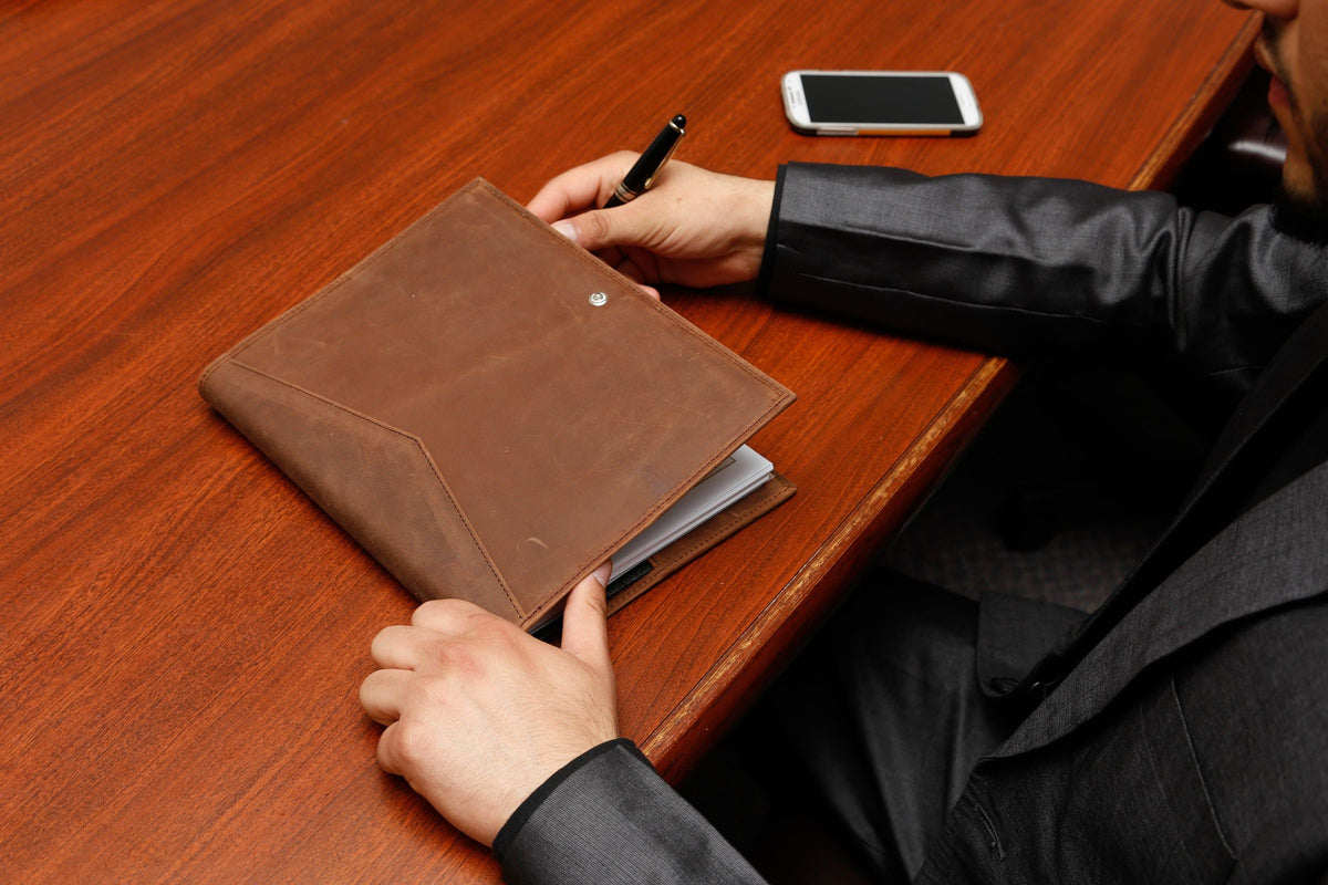 An executive in a suit writing notes in a brown leather folder at a wood conference table