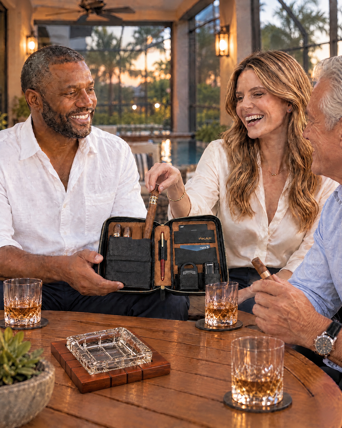 Three lawyer friends on a screened Florida lanai at dusk: the host holds the Xikar Sentinel Leather Cigar Case open while a laughing woman draws an unlit cigar from its main pocket and a silver-haired friend holds the cigar he has already taken; a wood and crystal cross-hatched ashtray sits on the teak table