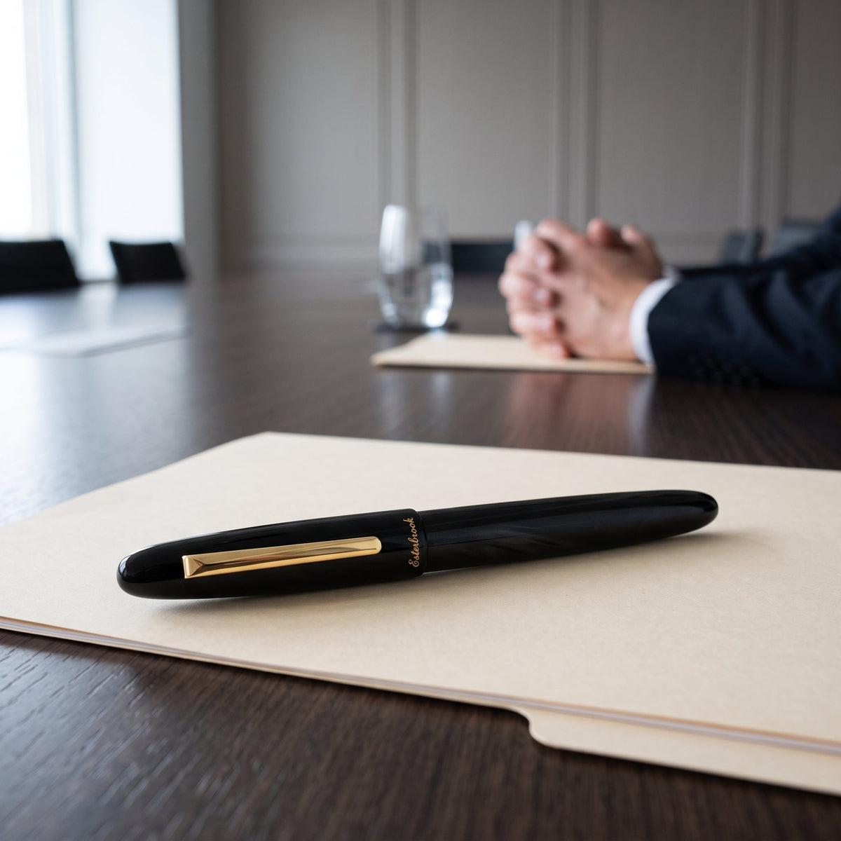 The Esterbrook Estie Rollerball in Ebony lying capped on a closed buff folder at a dark conference table, the folded hands and dark suit of a second person out of focus across the table