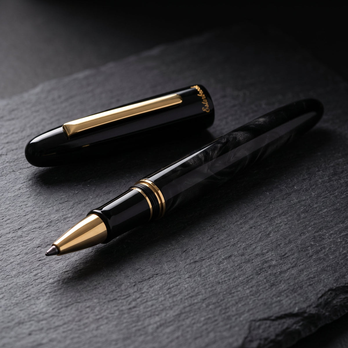 The Esterbrook Estie Rollerball in Ebony uncapped on dark slate in raking light, cap resting behind it, showing the figure in the black blended acrylic, the flat gold clip, the gold section bands and the gold rollerball cone