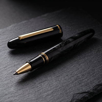 The Esterbrook Estie Rollerball in Ebony uncapped on dark slate in raking light, cap resting behind it, showing the figure in the black blended acrylic, the flat gold clip, the gold section bands and the gold rollerball cone
