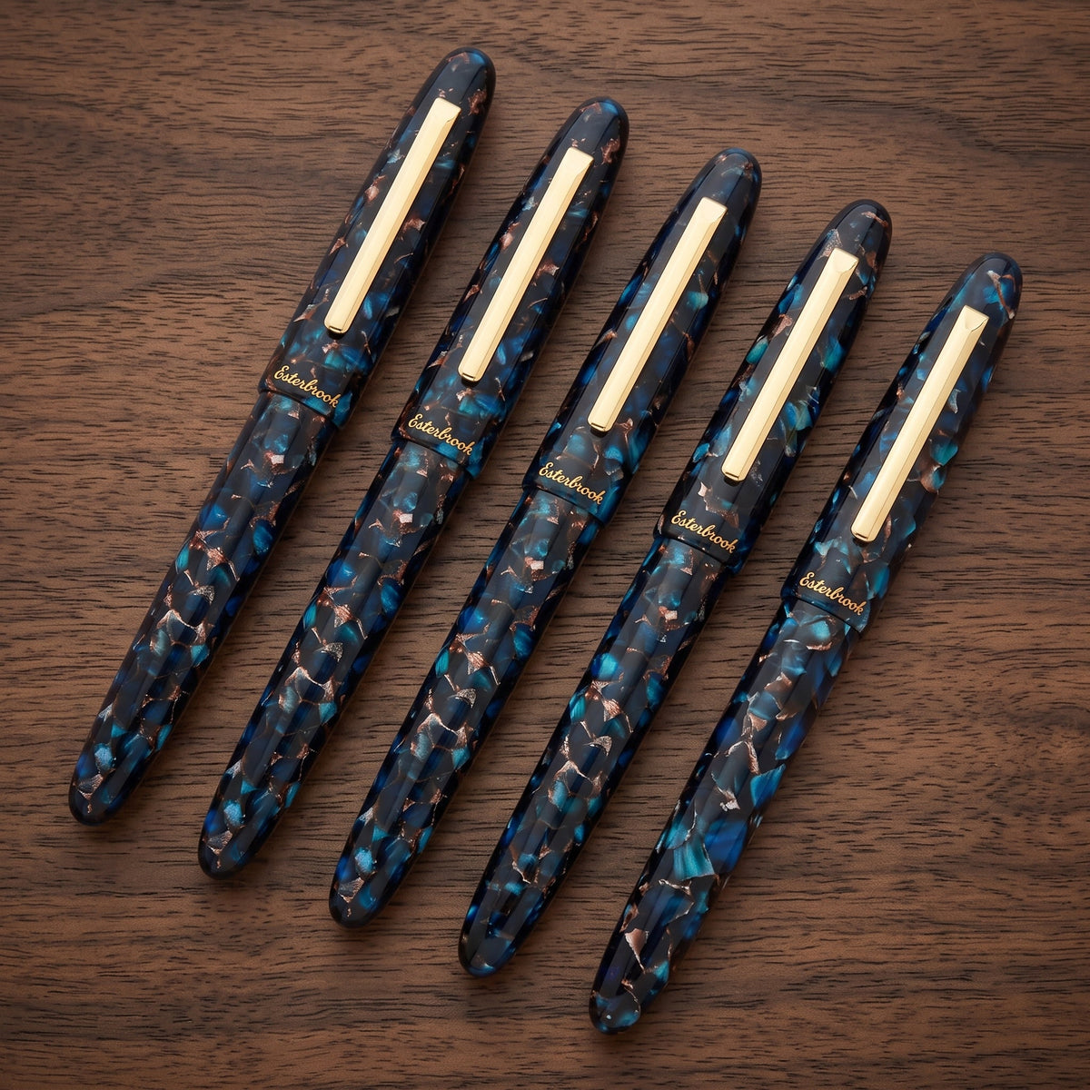 Five Esterbrook Estie Rollerballs in Nouveau Bleu laid capped in a row on a walnut table, clips aligned and each cap marked Esterbrook — identical across a run, as a firm would order for a class of associates