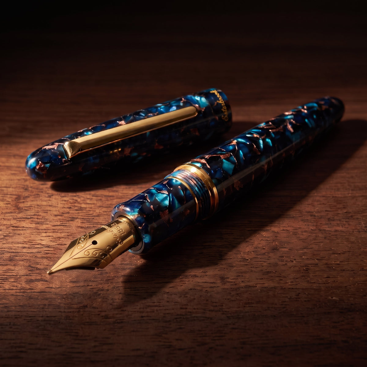 The Esterbrook Estie fountain pen in Nouveau Bleu uncapped on a dark walnut desk in raking light, cap resting behind it, showing the mosaic acrylic barrel, the flat gold clip and the solid 18-carat gold nib