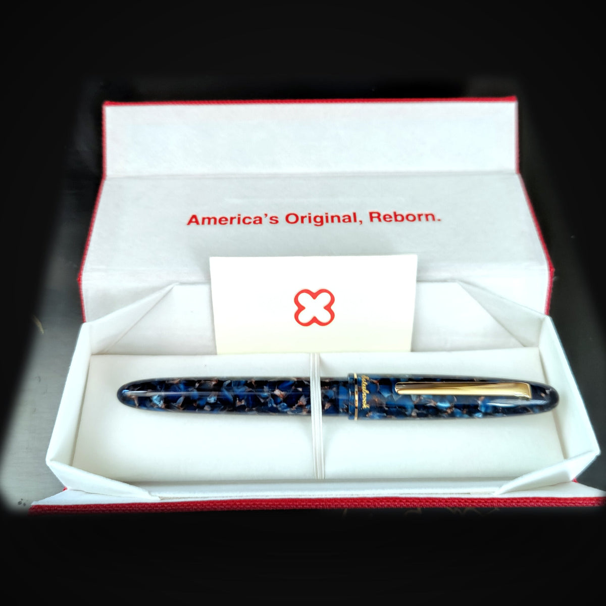 The Esterbrook Estie Rollerball in Nouveau Bleu capped in its gift box — red woven fabric outside, white lining printed America's Original, Reborn., the pen held under an elastic band beside a card bearing the red Esterbrook clover