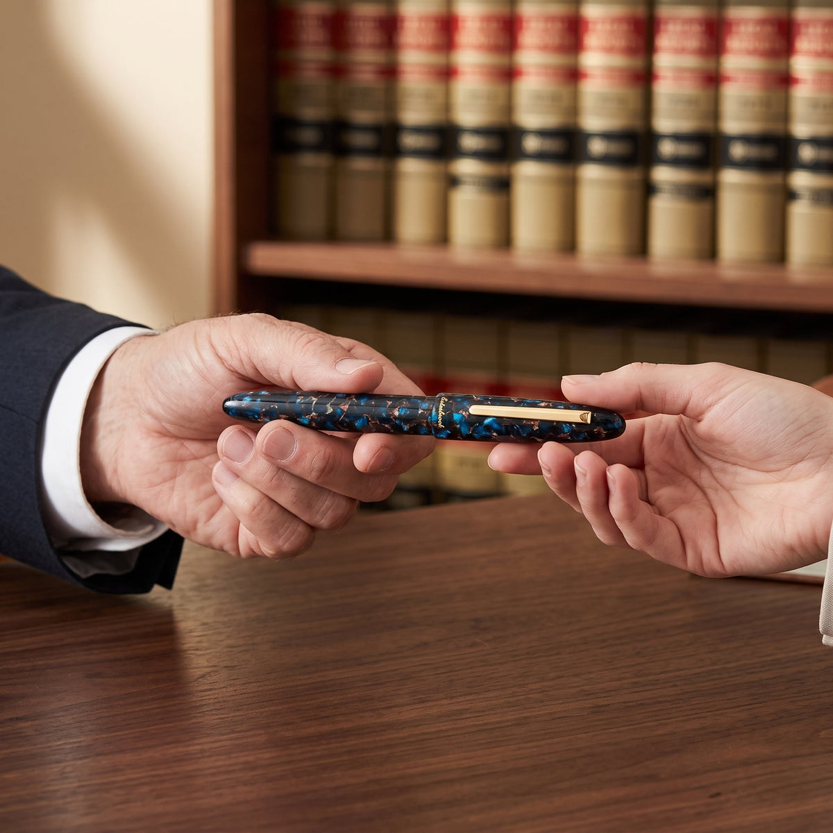 An older attorney hands the capped Esterbrook Estie fountain pen in Nouveau Bleu across a walnut desk to a younger colleague, a wall of law reports behind them