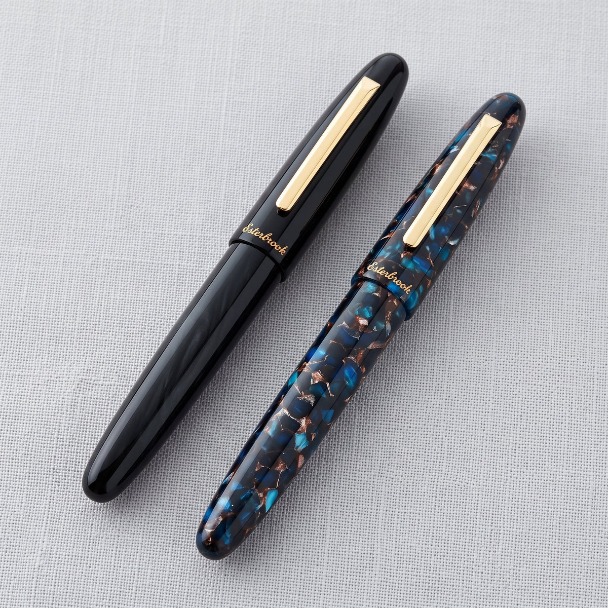 The Esterbrook Estie Rollerball in Ebony, at left, shown capped beside the Nouveau Bleu on pale grey linen — gloss black against navy, turquoise and copper mosaic acrylic, for comparing the two finishes