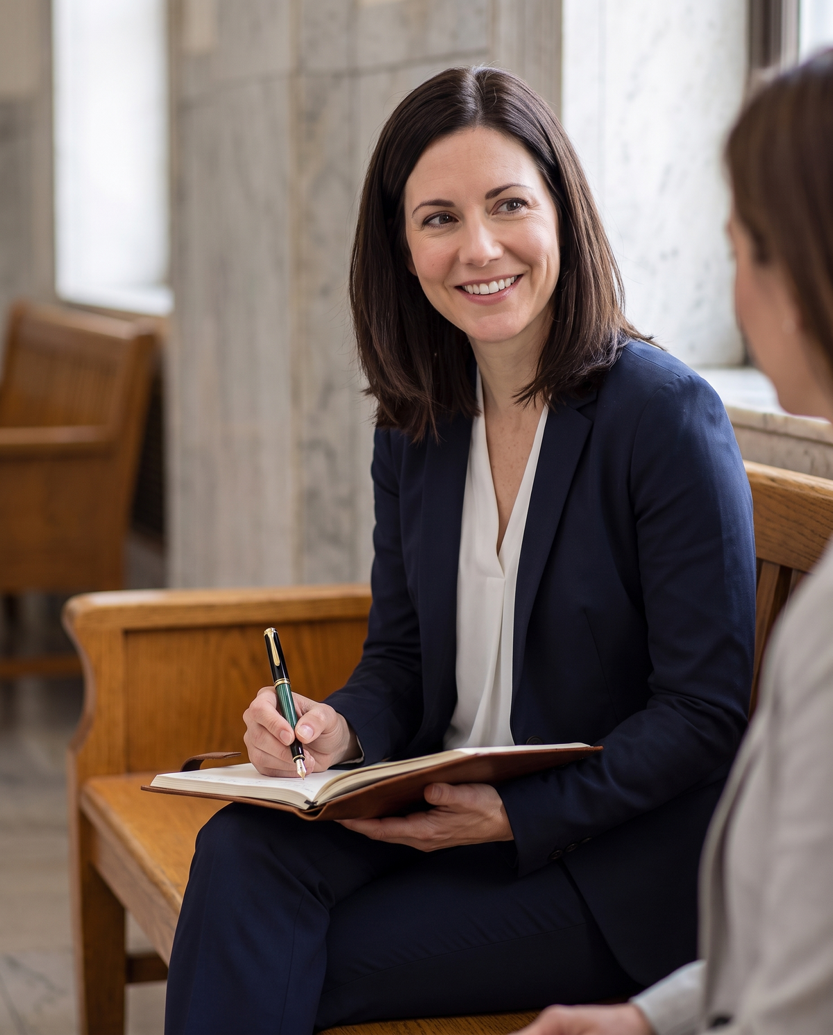 Pelikan Souverän M400 Fountain Pen — Black-Green: a woman lawyer in a navy suit writes in a leather notebook on a courthouse corridor bench, smiling at her client