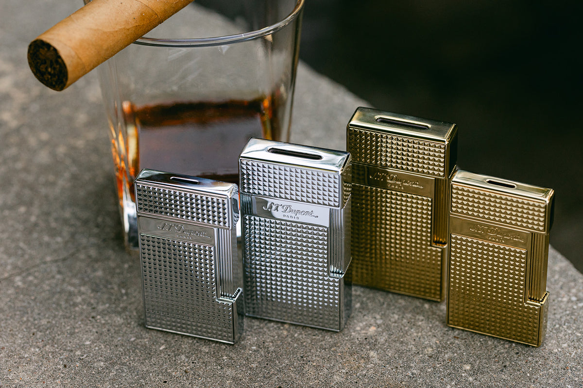 Four S.T. Dupont lighters in chrome and gold lined up by height beside a cigar and a glass of whisky