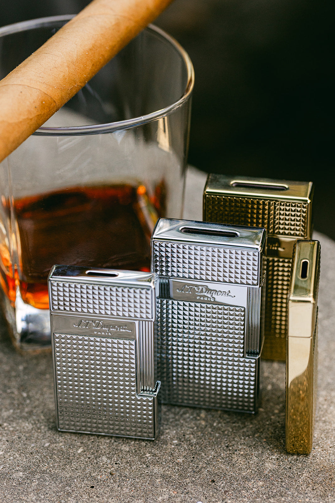 Four S.T. Dupont Slimmy and Biggy lighters in chrome and gold grouped beside a cigar and a glass of whisky