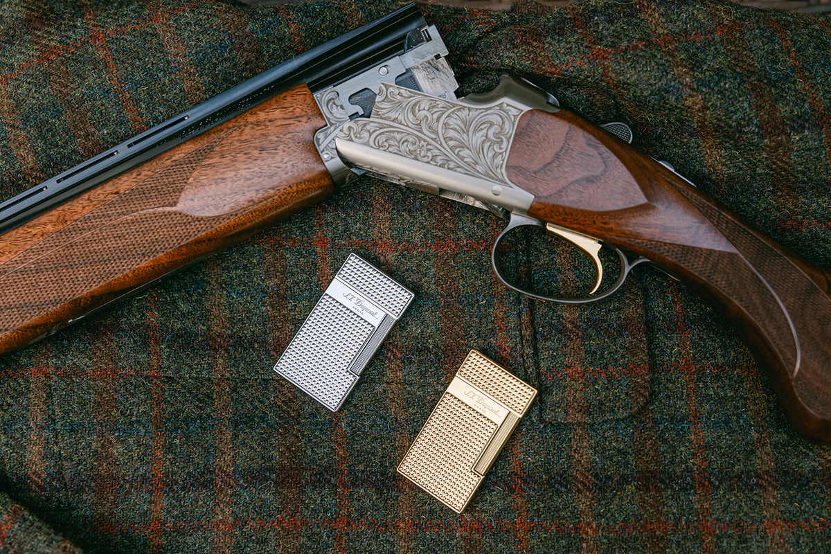 Chrome and gold S.T. Dupont Diamond Head lighters on a tweed blanket beside an engraved over-and-under shotgun