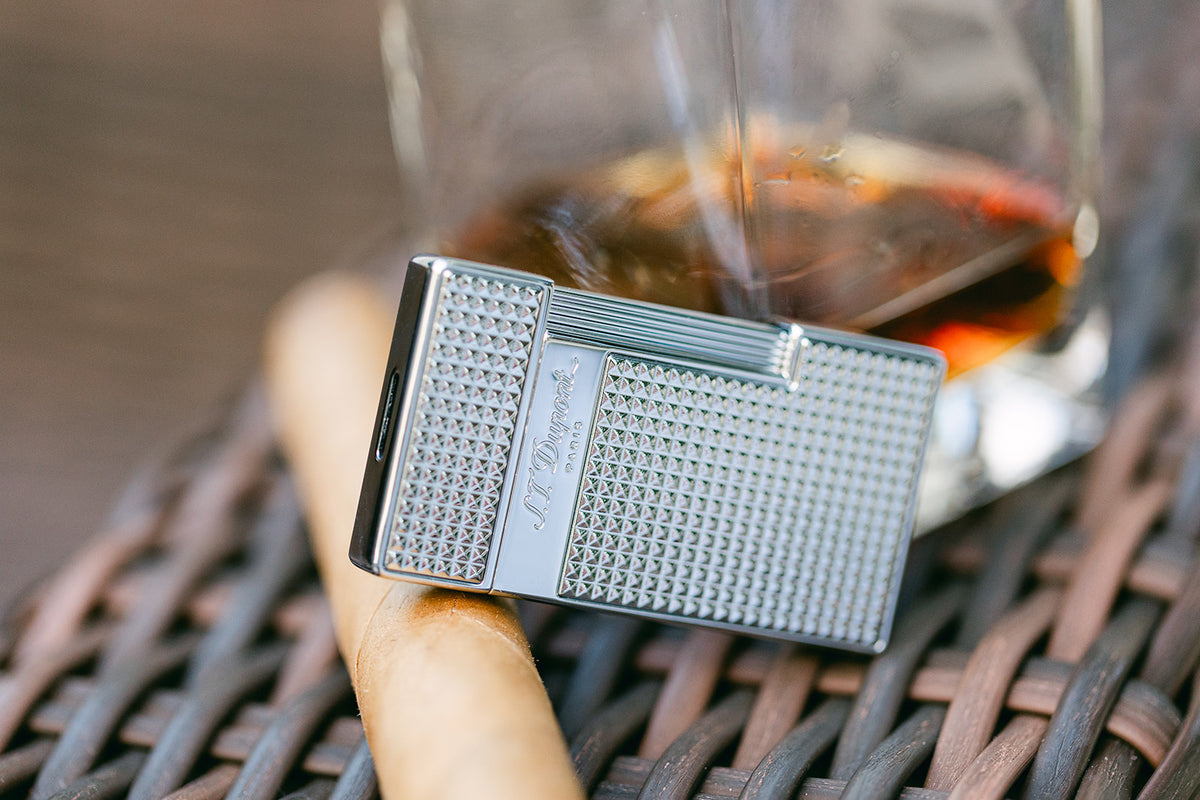 The chrome S.T. Dupont Slimmy lighter resting on a cigar beside a glass of whisky on a wicker table